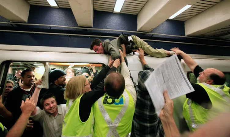 Photo of crowds of people at a BART station, including protesters grasping the roof of the train car, being pulled down by people in safety vests.