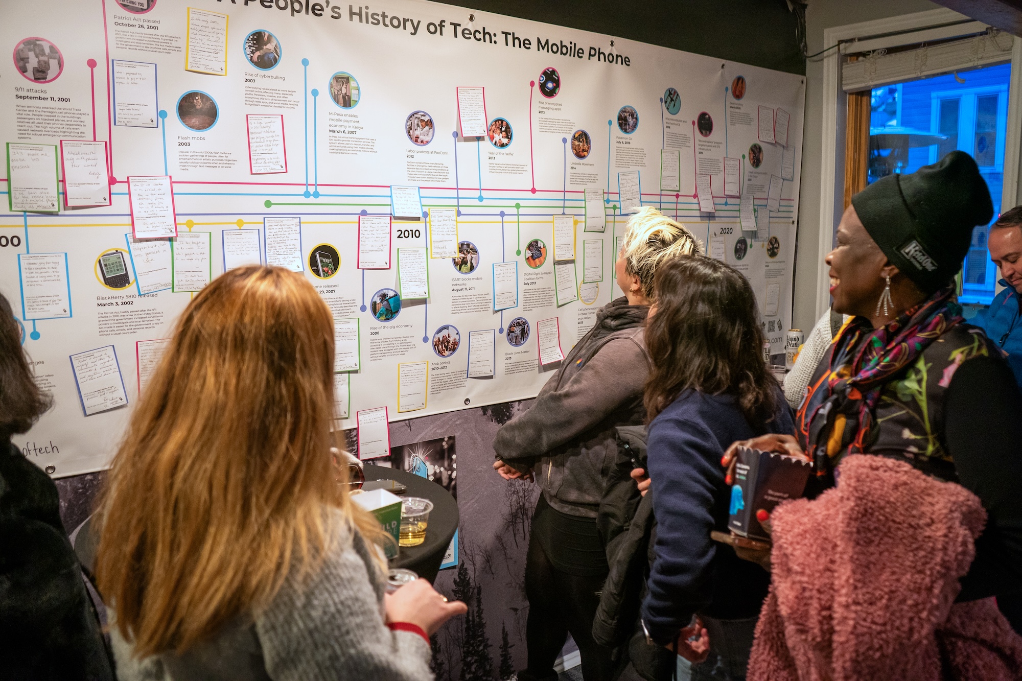 A crowd of participants gathers around a timeline banner covered in post-it notes of handwritten personal contributions to the timeline.